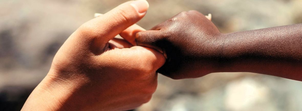 Hand eines afrikanischen Kindes greift den Zeigefinger einer weißen Hand, symbolisiert die weltweiten Hilfsprojekte der Kamillianer
